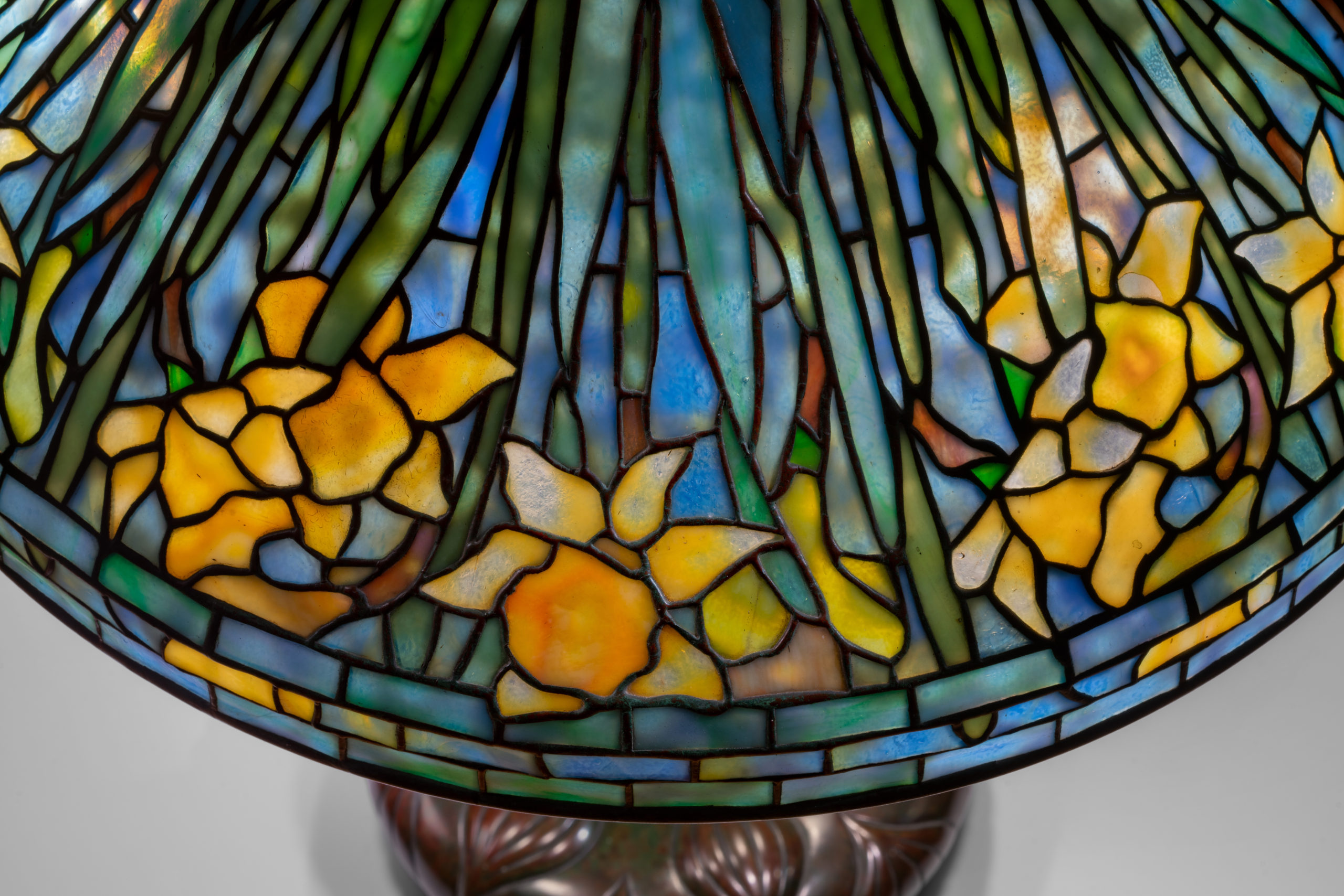 A close up view of a blue and green mosaic glass lampshade accented with a yellow daffodil repeated pattern around the rim.