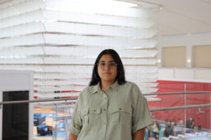 A Colombian-American woman with dark brown long hair, silver glasses, and silver hoop earrings wearing a green and white striped shirt stands in in front of a glass railing at the Queens Museum.