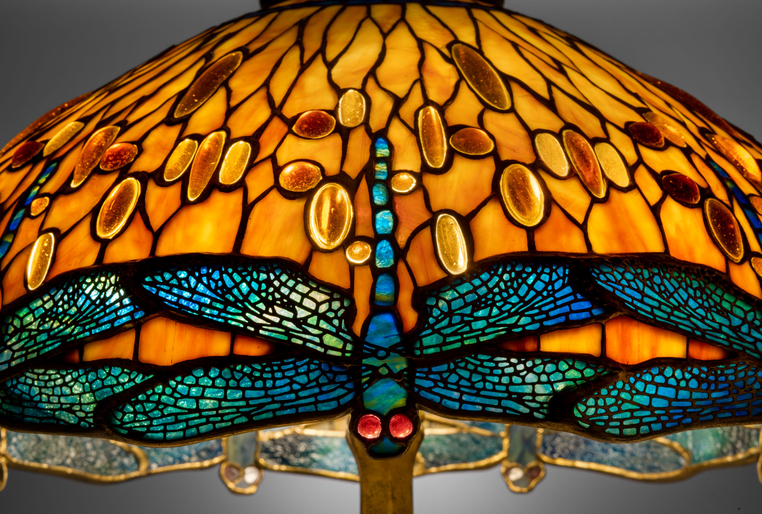 A close up view of an orange, glass, mosaic lampshade. The rim of the lampshade is made up of a repeated, upside down, blue dragonfly pattern. Some of the glass pieces have a jewel tone quality and are reflecting light.