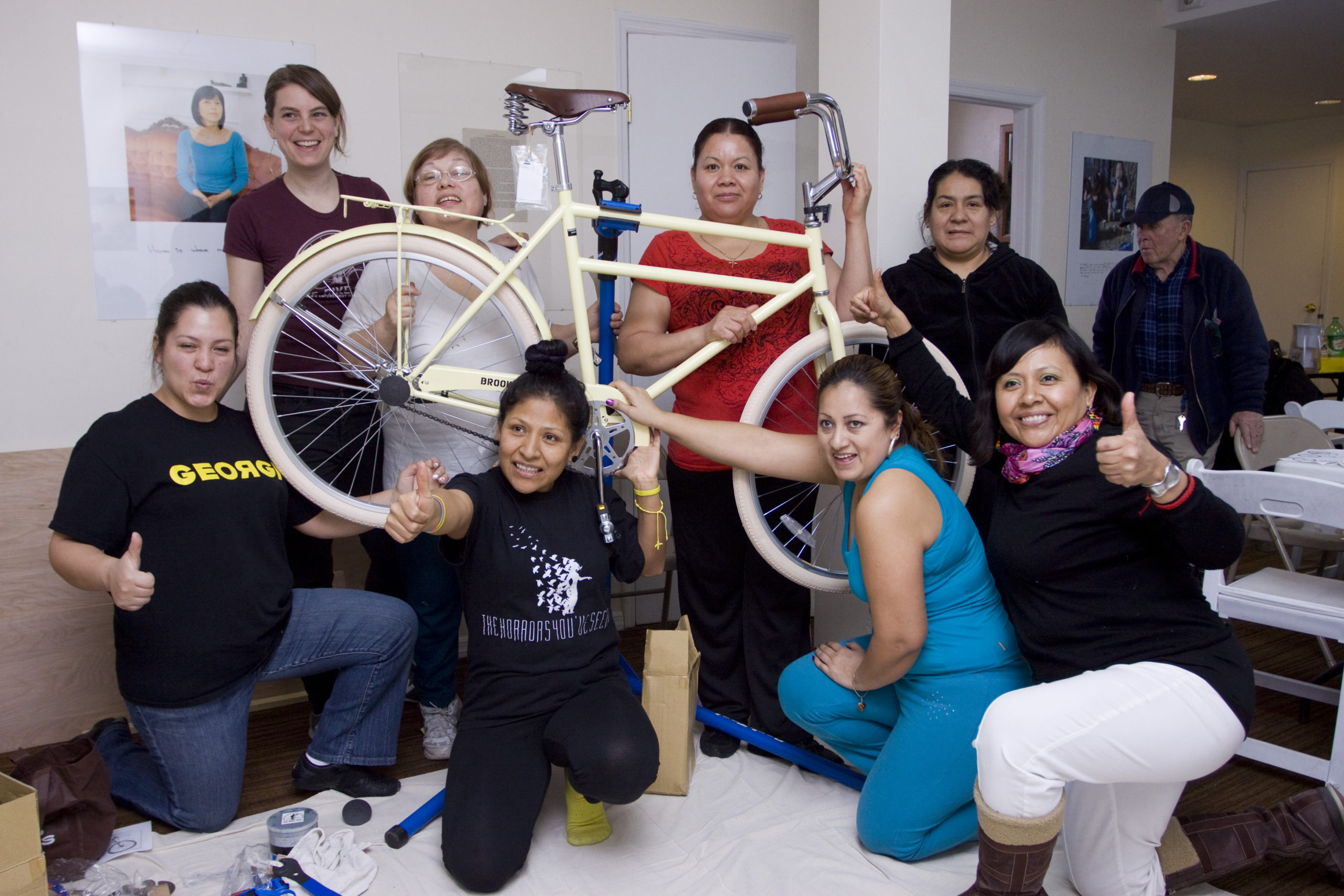A group of women pose for a photo in a workshop space. They are gathered around, and holding up, a cream colored bike. In the background are white walls displaying art and an elderly man standing beside a table with folding chairs.