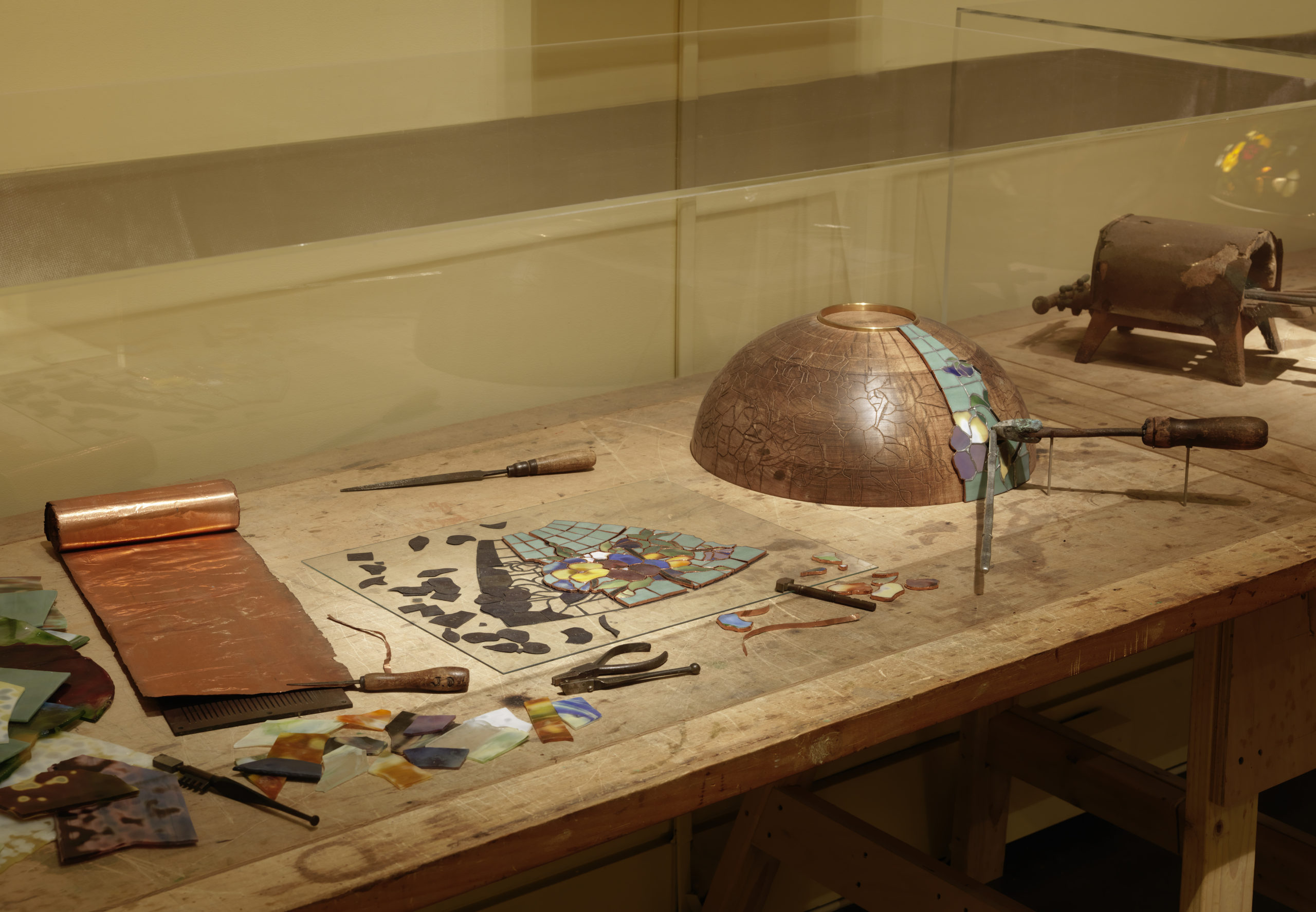 A display case showcasing tools used to fabricate Tiffany Glass lamps. On the display table lie a selection of metal and wooden tools, sheets of copper, shards of colorful glass, and a copper half-sphere mold demonstrating how Tiffany Glass lamps are assembled.