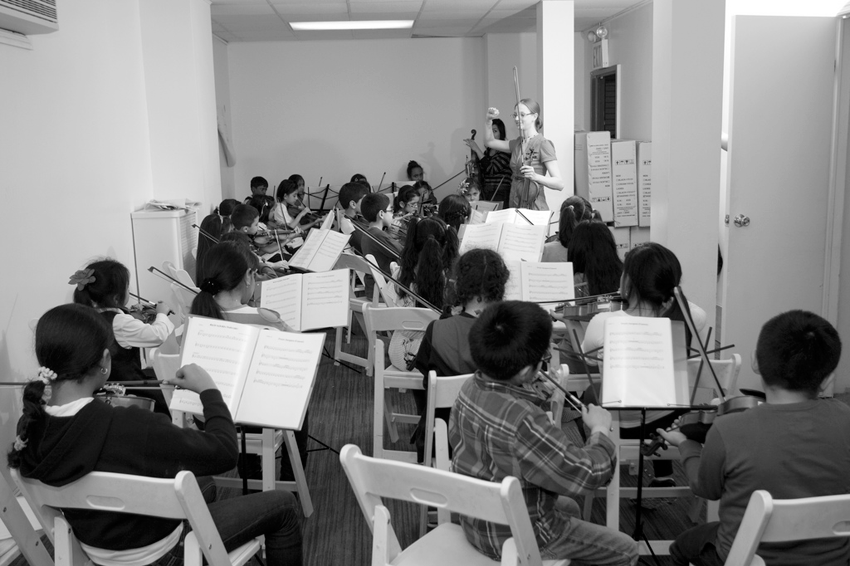 A black and white photo of a classroom, filled with Latinx children seated in folding chairs. They are all viewing booklets of sheet music in pairs, and practicing playing the violin. In the far right corner are two adult instructors.