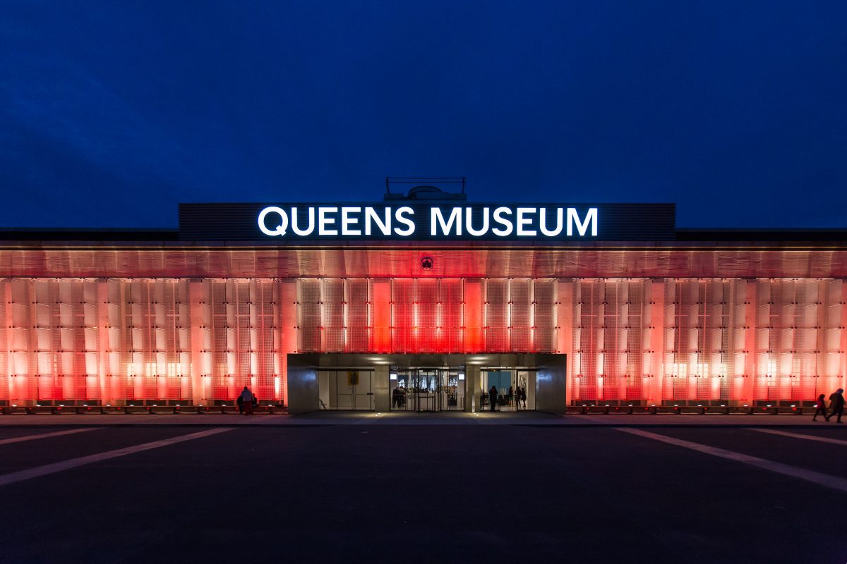 A landscape view of the entrance to the Queens Museum, against a dark blue evening sky. The museum is glowing red and in the top center of the building, in white uppercase font, reads “QUEENS MUSEUM”.