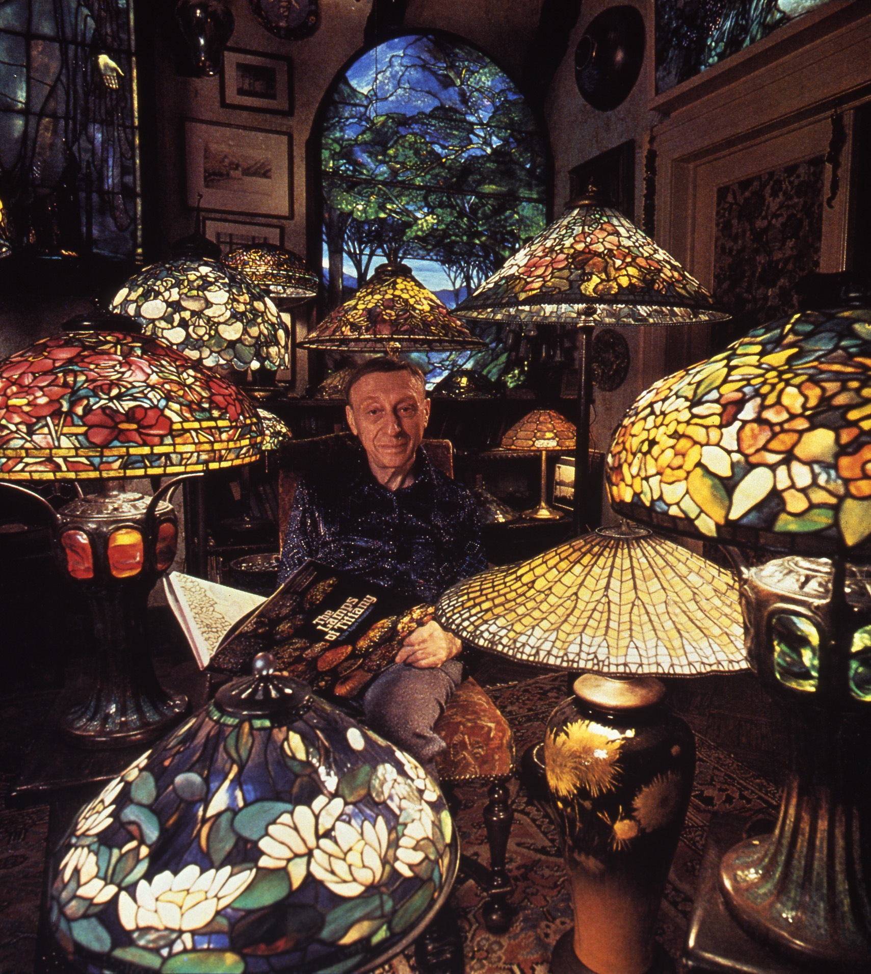 Dr. Egon Neustadt sitting in a brown, cushioned chair. He is wearing a blue button up and gray slacks. His face is framed and lit up by a sea of glass lamps with different, colorful, mosaic patterns. Behind him is a large, blue and green, stain-glass window of a nature scene.