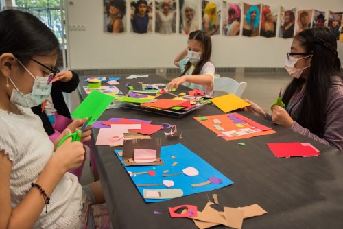 Three young children wearing facemasks, seated at a table with bright colored paper, cutting and collaging creating artwork.