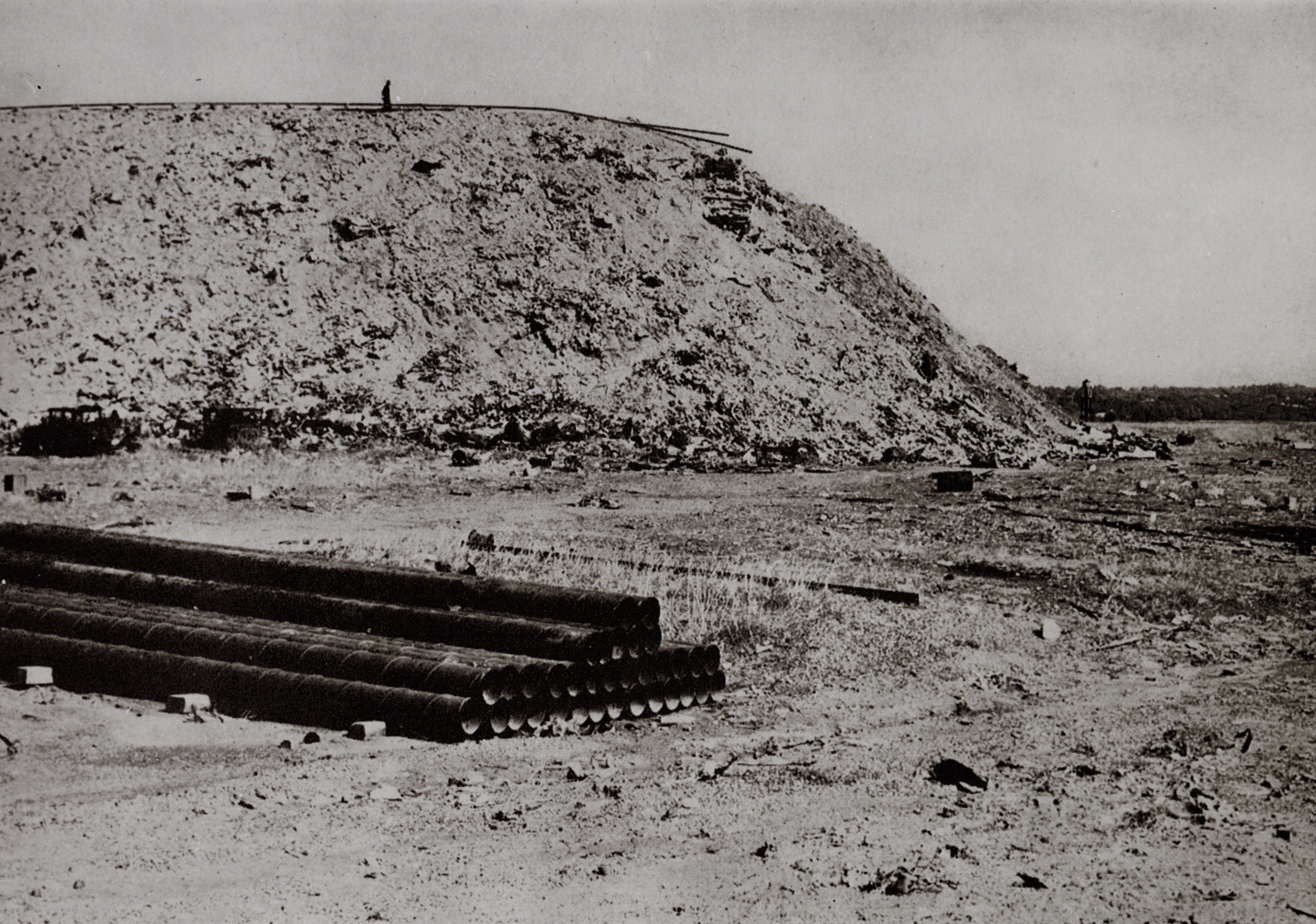 A black and white photo of a meadow, with a 90 feet tall mound of burnt coal ash. At the foot of the mound lies a stack of cylindrical pipes. On top of the mound is a silhouette of an adult.