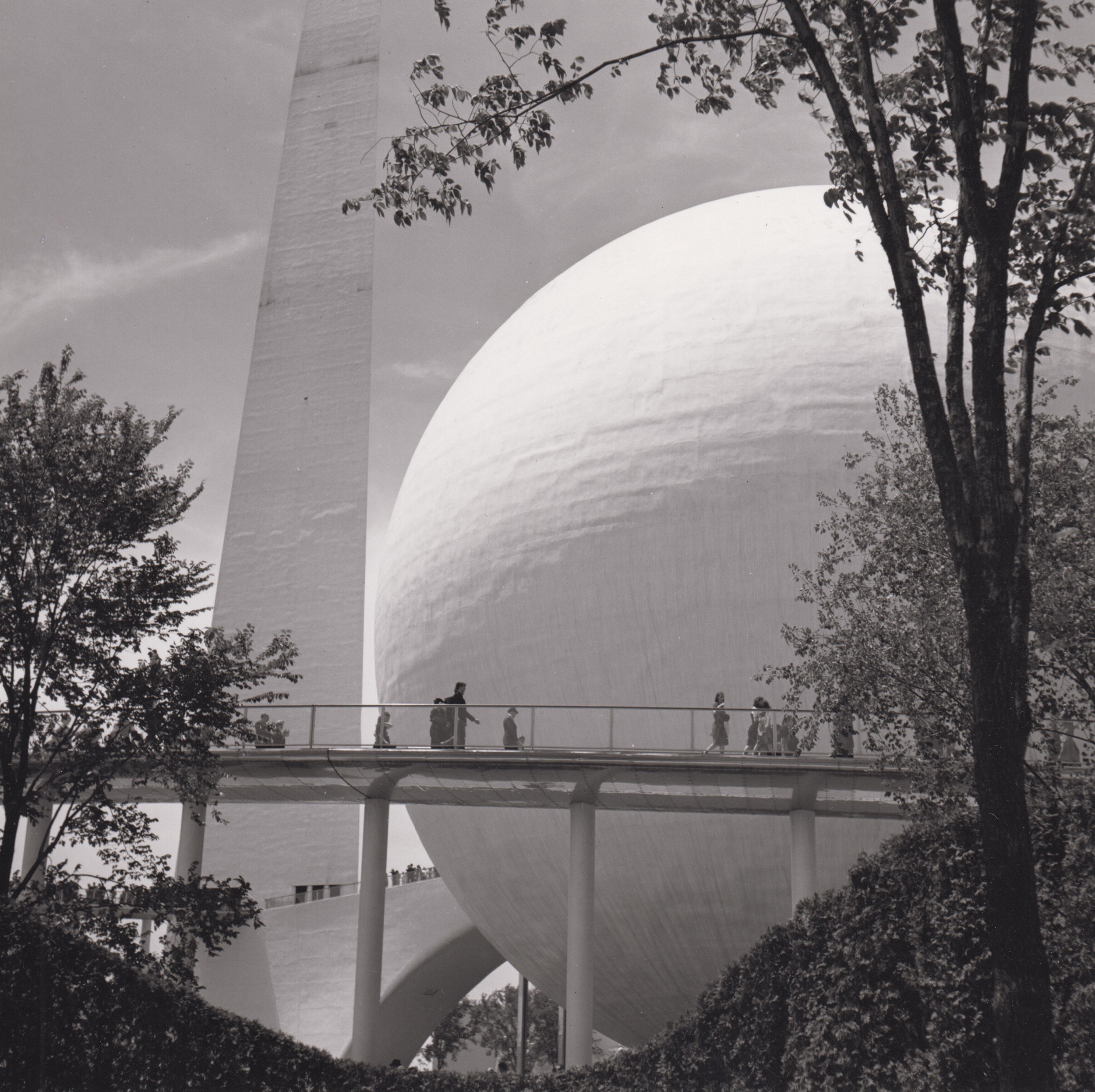 A black and white photo of two grand, modern sculptures with a triangular and spherical shape. With confident presence they occupy a sky with wispy clouds. In front of the sculptures is a raised walkway with people walking by. Framing the view is foliage and trees. 