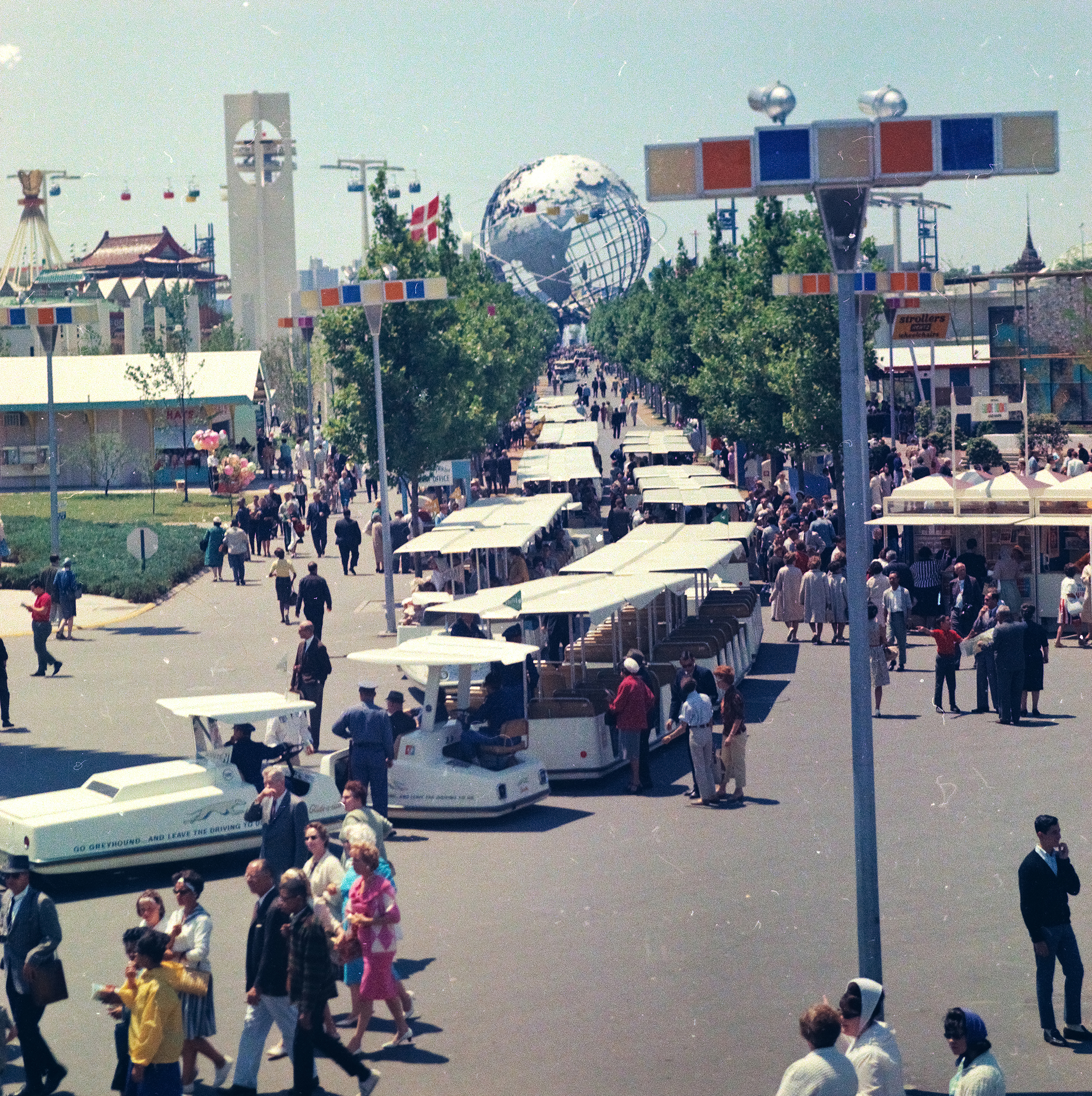 A photo of a pavilion avenue filled with open sightseeing shuttle buses during the 1964-65 World's Fair. The avenue is filled with patrons and lined with happy green trees that lead down to the Unisphere. 