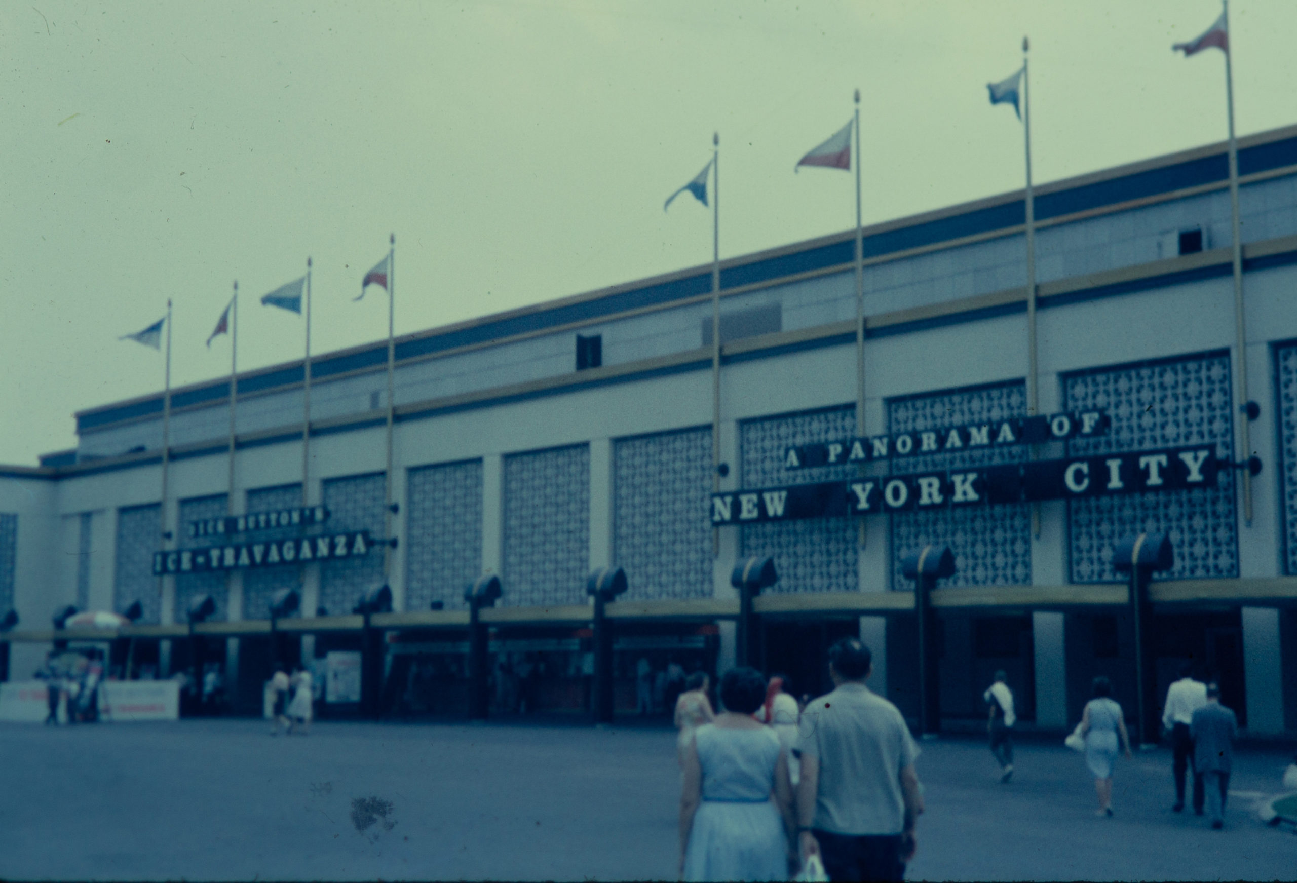 A dim, colored film photo of the New York City Building. Adults are dispersed in the walkway in front of the building. The building is lined with flags and there are partial views of the building’s signs. The one on the left reads “A PANORAMA OF NEW YORK CITY” in uppercase font.