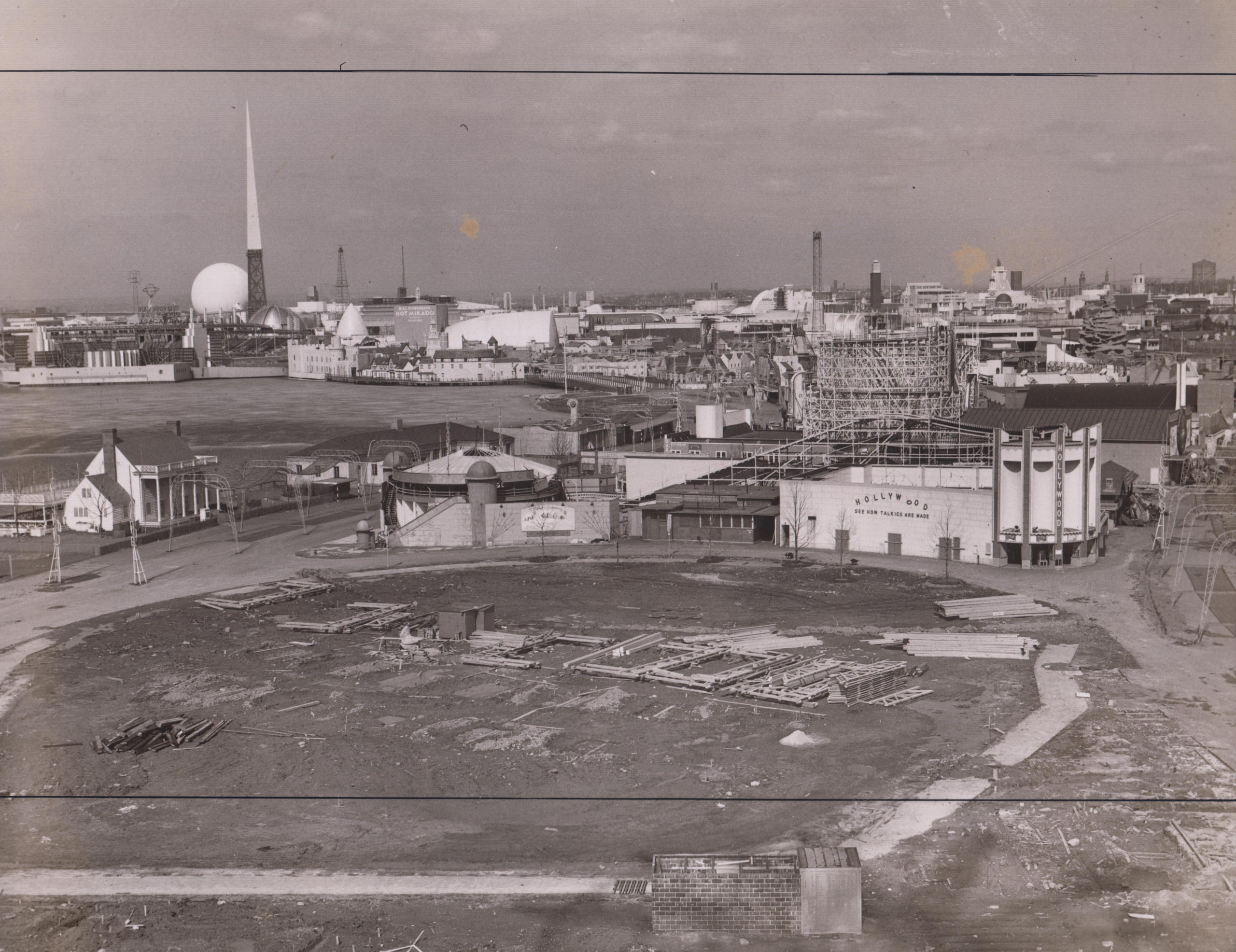 A black and white photo of an open piece of flat land. Sitting on the top of the land is a row of building material stacked in the center. Behind that, is a configuration of buildings of different types. The nearest building has a sign on the side that reads “HOLLYWOOD, SEE HOW TALKIES ARE MADE”.