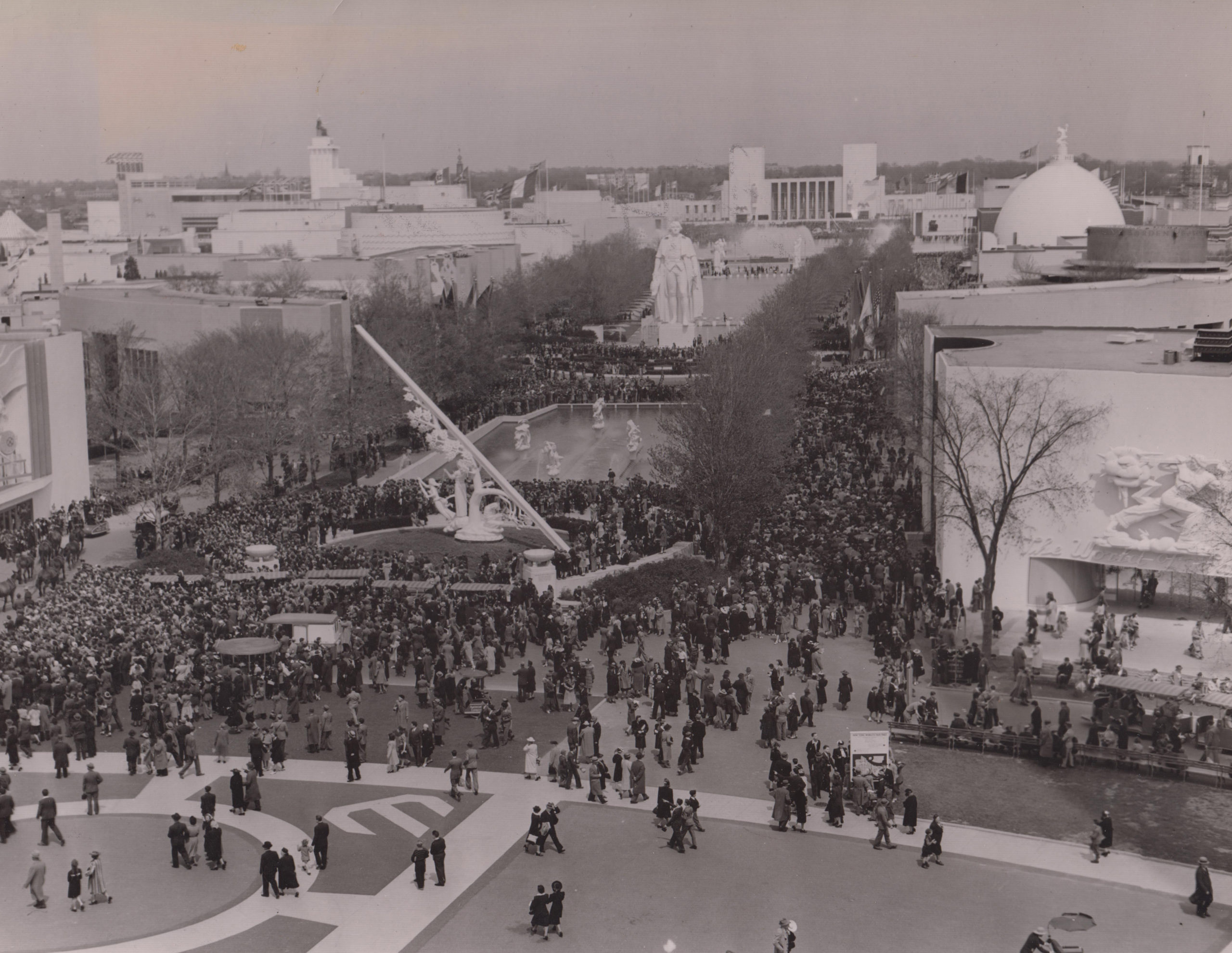 A black and white photograph of an upside down t-shaped plaza. A sea of patrons pack the streets. In the center aisle are monuments and fountains. The plaza is lined by two rows of trees and pavilions. 