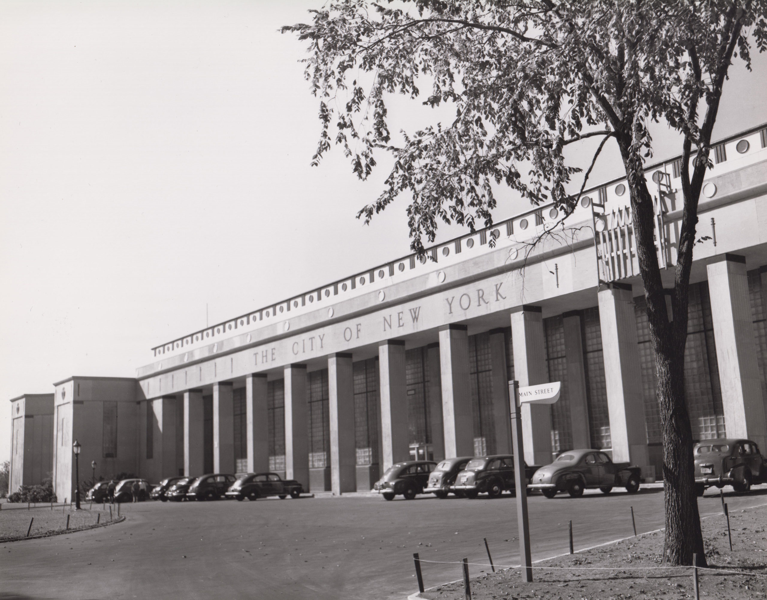 A black and white photo of the New York City Building. It has a long rectangular shape with a row of square columns. At the top of the building in uppercase font reads “THE CITY OF NEW YORK”. In the parking lot, in front of the building, are nine cars parked in a row and a street sign, to the right, that reads “MAIN STREET '' in uppercase font.