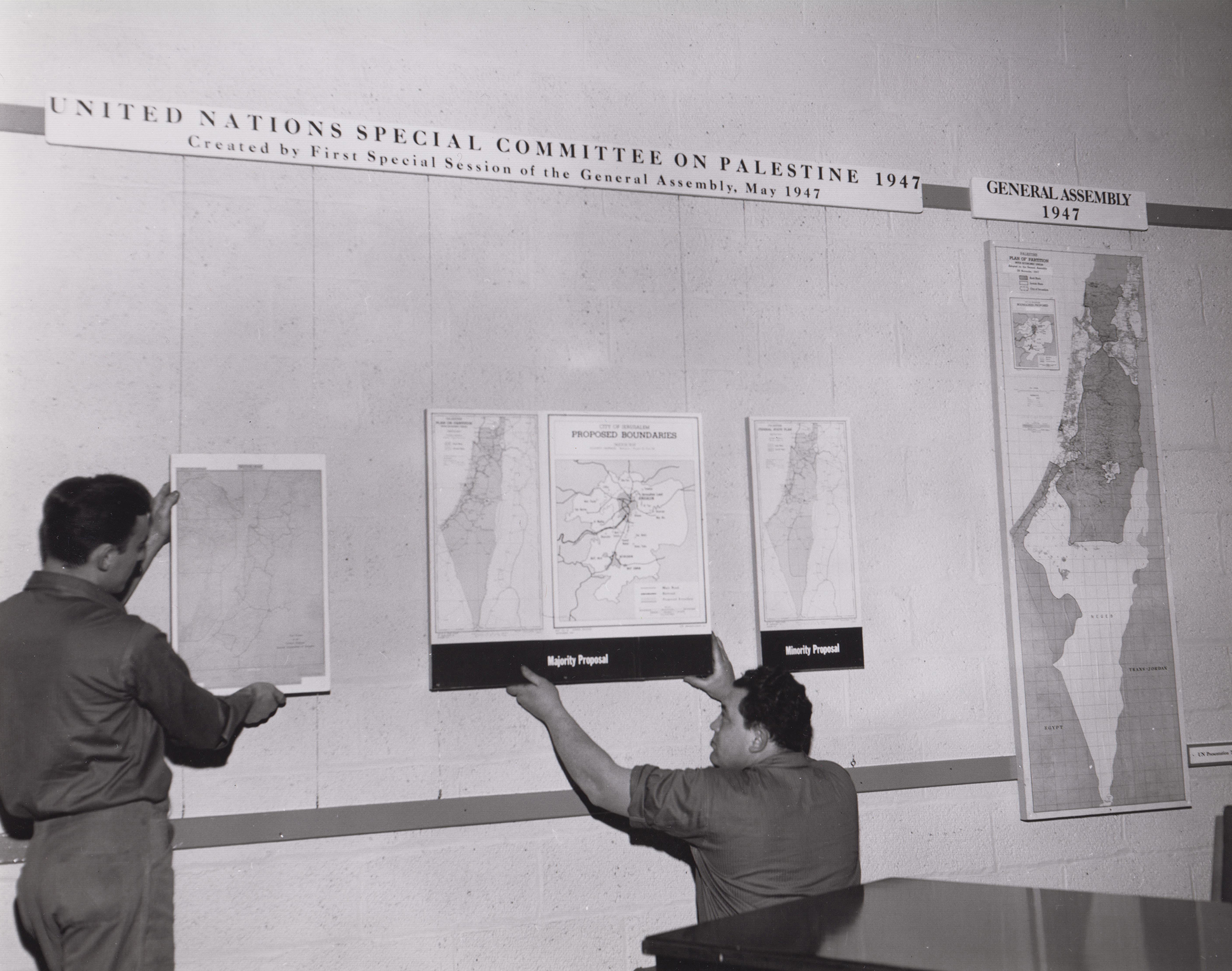 A black and white photo of two men installing boundary maps of Palestine. One is sitting and one is kneeling as they access the placement. Below the center trio of maps are two labels that read “Majority Proposal” and “Minority Proposal”. Above the row of maps is a portion of a timeline. The timeline reads "UNITED NATIONS SPECIAL COMMITTEE ON PALESTINE 1947, Created by First Special Session of the General Assembly, May 1947” and “GENERAL ASSEMBLY 1947”.