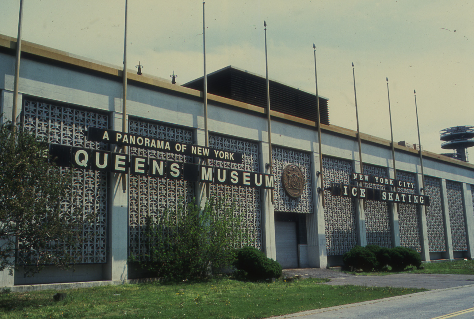 A colored film photo of the Queens Museum against a cloudy sky. The building is white with an intricate pattern on the facade. There are two sets of black building signs that read “A PANORAMA OF NEW YORK, QUEENS MUSEUM” and NEW YORK CITY ICE SKATING” in white, uppercase font.
