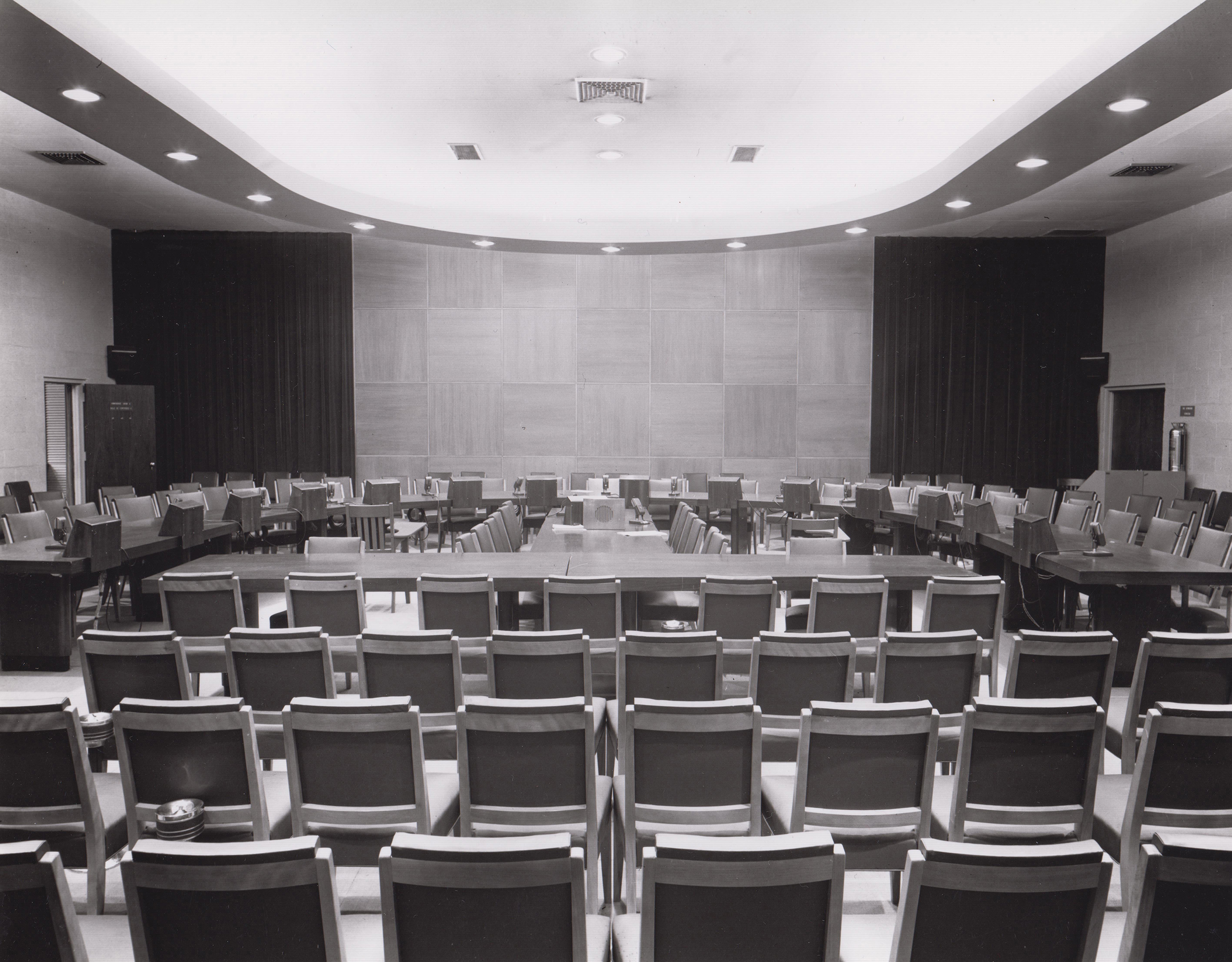 A black and white photo of an empty conference room. In the foreground are four rows of chairs. In the center are two tables placed perpendicularly. At the head of one of the tables, is a speaker. Completing the room is a group of tables, lined with chairs, arranged in a half circle. In the background are two exits and a set of open curtains. 