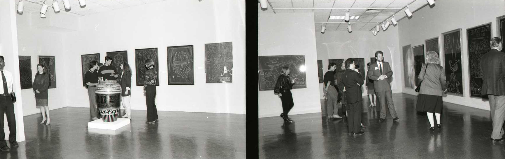Two black and white photos, side by side. Both share views of the same gallery. The photo on the right captures a security guard, six patrons, and a large, patterned vase installed on a podium, in the center of the floor. Keith Haring’s drawings, on black paper, are installed in a centered row on the walls. The photo on the right captures a similar scene. Drawings installed in the same fashion and patrons walking about the gallery space. 