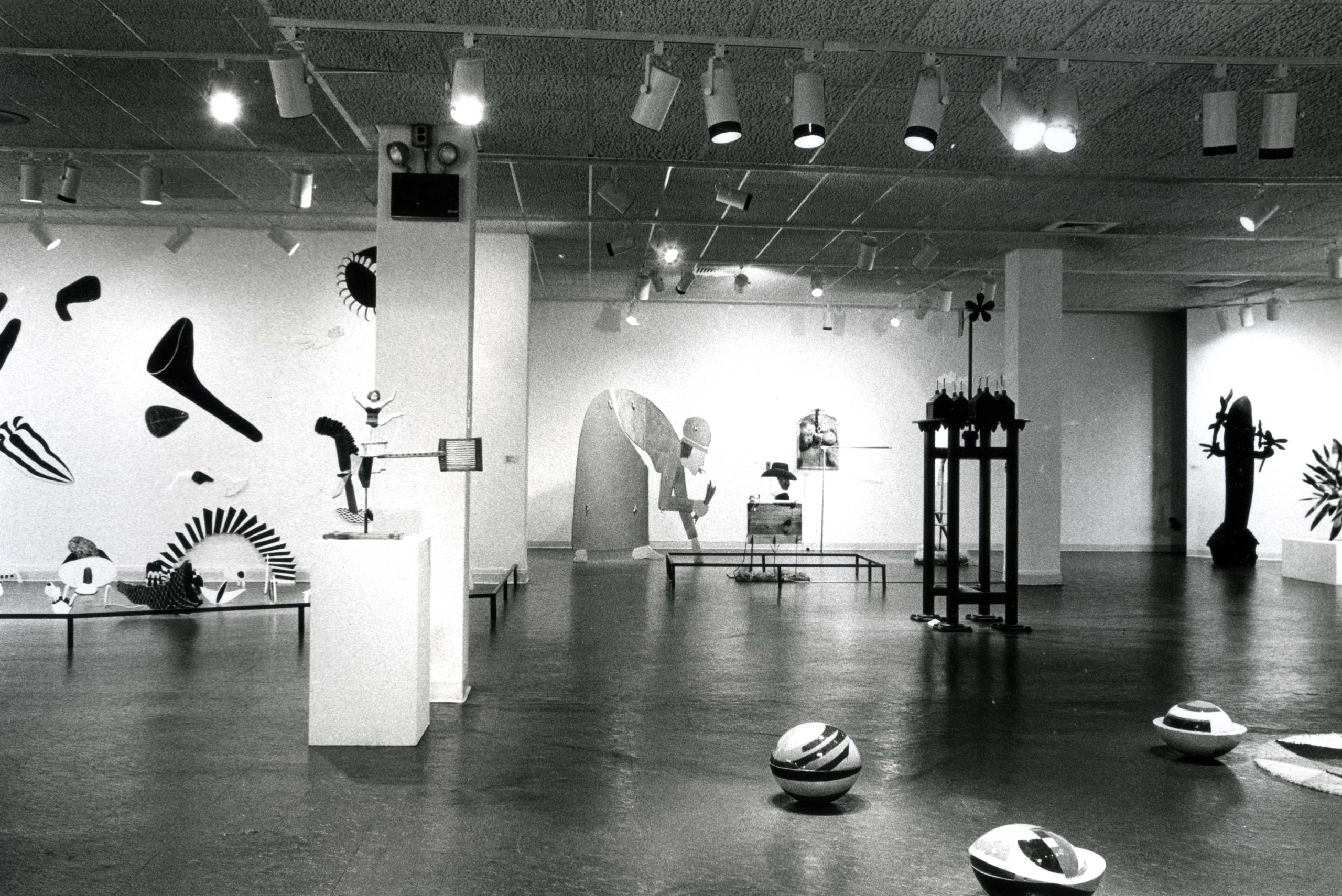 A black and white photo of an exhibition space filled with an eclectic mix of sculptures. Some are free standing, some are displayed on podiums and some are hanging on the back wall. 