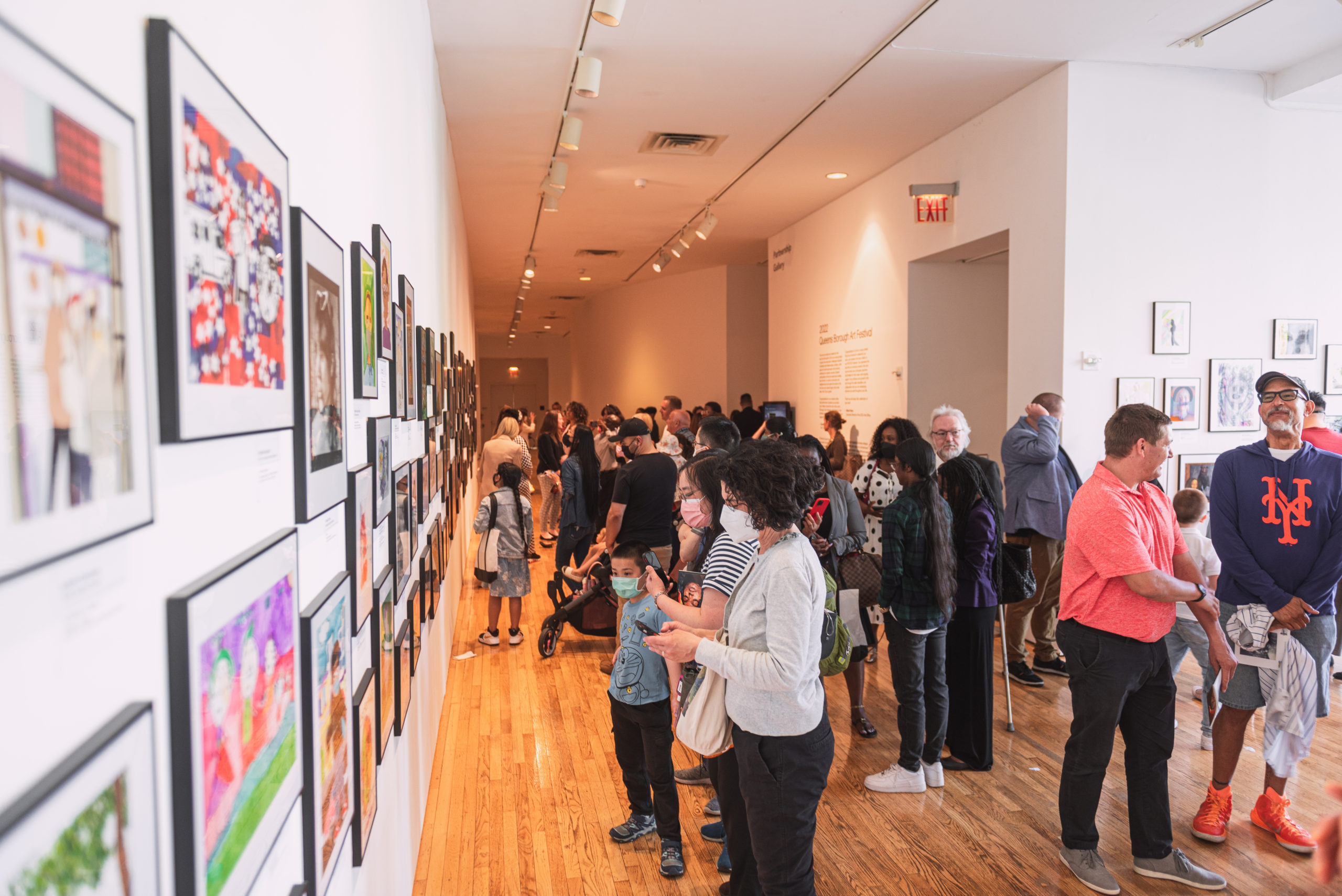 A narrow walkway in an exhibition space, filled with visitors of different races and ages. On the left is a wall full of framed artworks, installed salon style. 