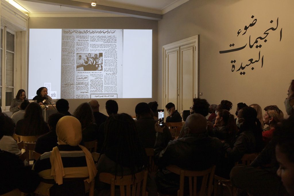 Nsenga Knight, a brown skinned Afro-Caribbean American woman wears a black islamic headscarf and stands in front of a microphone next to Samah Gafar, a brown-skinned, straight haired sudanese woman who is her arabic translator. She speaks into a microphone and behind her is a projection of a newspaper article written in arabic with a photograph of Malcolm X seated with his family. Knight is in front of a large seated indoor crowd at the Contemporary Image Collective, an arts institution in Cairo Egypt. There is Arabic writing on the wall. 