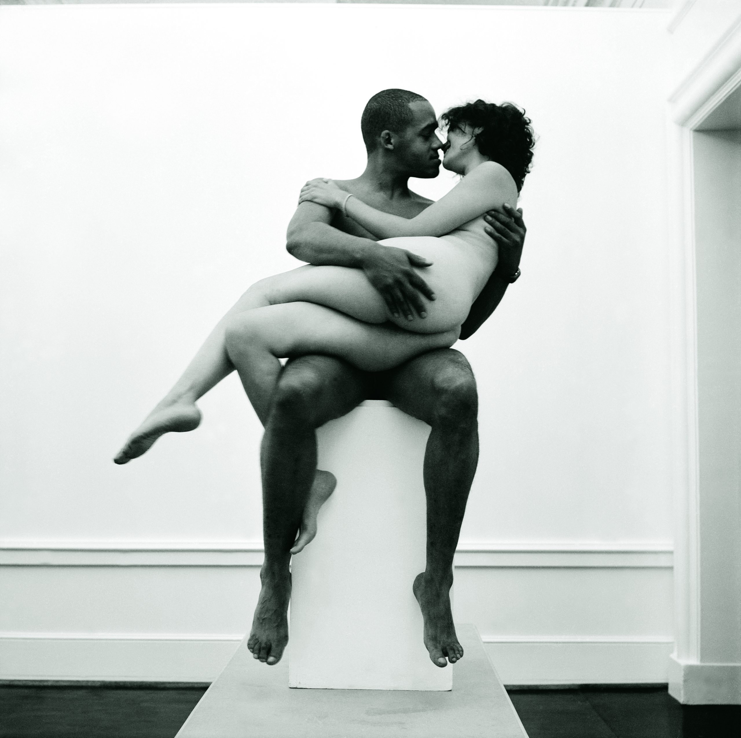 Black and white photograph depicting a light-skinned nude woman sitting on the lap of a nude brown man on a pedestal inside a gallery-like setting, reminiscent of a classical sculpture.