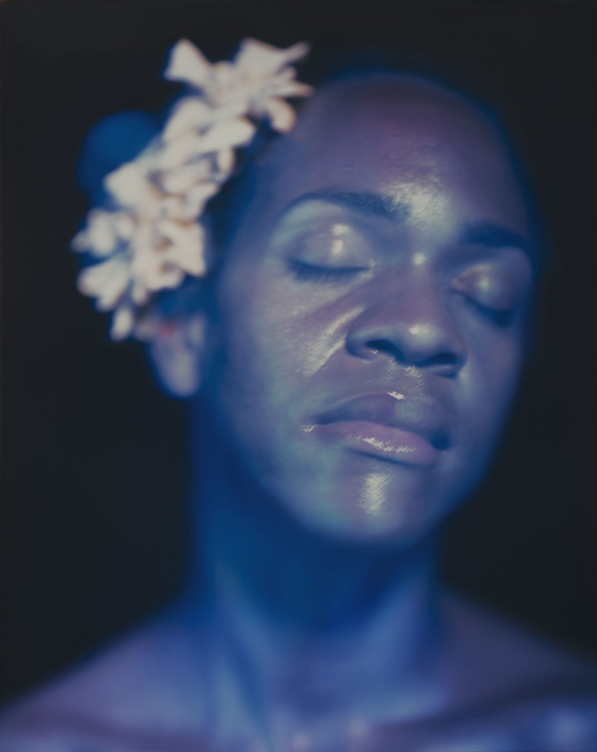 A tight headshot of artist Lyle Ashton Harris resembling singer Billie Holiday. His eyes are closed and his glowing skin is bathed in blue hues. A cluster of flowers are pinned to the side of his head.