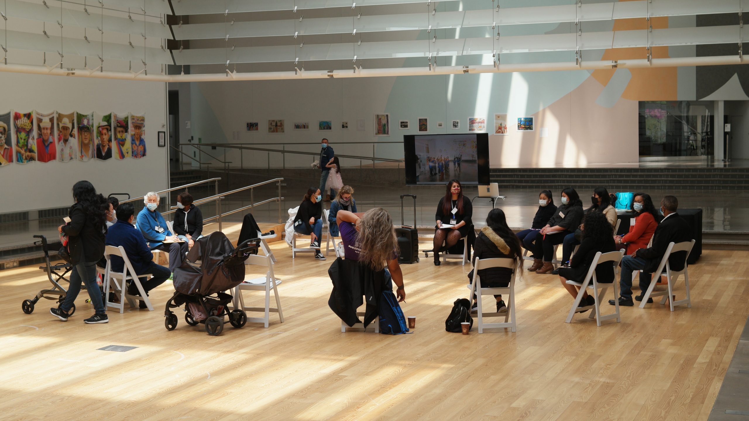 A photograph taken in mid-day during a focus group held at Queens Museum, one of the participatory design activities that took place during the Central Atrium for All project. A group of about 20 individuals wearing surgical masks are seated in a circle of wooden folding chairs, awash in a golden glow of filtered daylight from a skylight above. The diverse group includes older adults, individuals with walkers, caregivers, baby strollers, and white canes for low-vision individuals. The group is in active discussion with a facilitator, who sits in front of an amplifier and large digital TV monitor that depicts children on a tour of the museum.